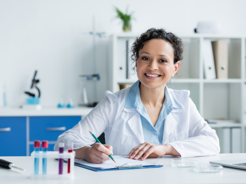 Causes of a UTI: A female scientist in a lab coat smiles while writing on a clipboard at a desk with test tubes and petri dishes, with laboratory equipment and shelves in the background.