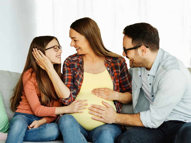 Can being pregnant cause a UTI? A smiling pregnant woman sits on a couch with a girl and a man. The girl touches the woman’s face lovingly, and the man has his hand on the woman’s belly. All three look happy and connected.