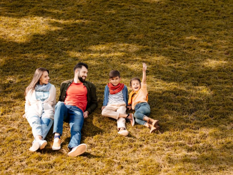 Can constipation cause a UTI? A family of four sits on the grass, smiling. Two adults are on the left, and two children are on the right, with one child raising their hand excitedly. The sun casts shadows on the grassy field.