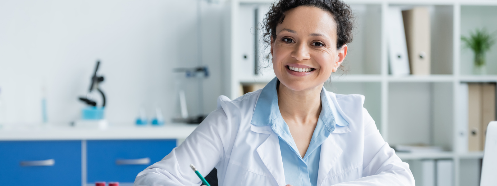Causes of a UTI: A smiling woman in a white lab coat sits at a desk with papers, holding a pen. Shelves with folders, a microscope, and lab equipment are visible in the background.