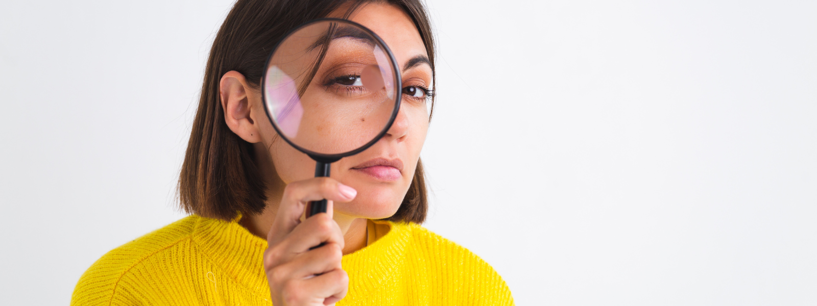 Can antibiotics cause a UTI? A person wearing a yellow sweater holds a magnifying glass to their face, enlarging one eye while looking directly at the camera, as if closely examining common UTI myths against a plain white background.