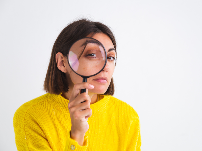 Can stress cause a UTI? A woman in a yellow sweater holds a magnifying glass to her face, playfully enlarging one eye as she looks for the truth behind UTI myths. She has straight, shoulder-length dark hair and stands against a plain white background.
