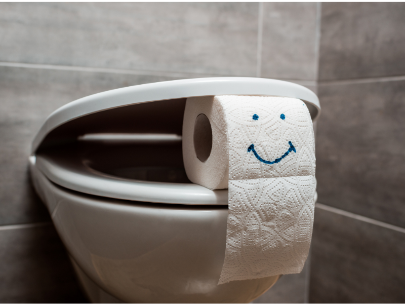Can you get a UTI from a toilet seat? A roll of toilet paper hangs on a closed toilet seat, with a smiling face drawn in blue on the front sheet. The background shows gray tiled walls.