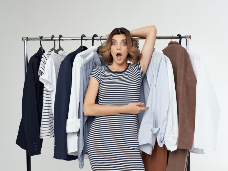Can your clothes cause a UTI? A surprised woman in a striped dress stands next to a clothes rack filled with various shirts and jackets, pointing at the clothes with her mouth open and one hand on her head.